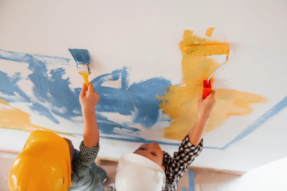 Dwóch malarzy w kaskach maluje sufit wałkami w niebieskim i żółtym – Malowanie sufitów