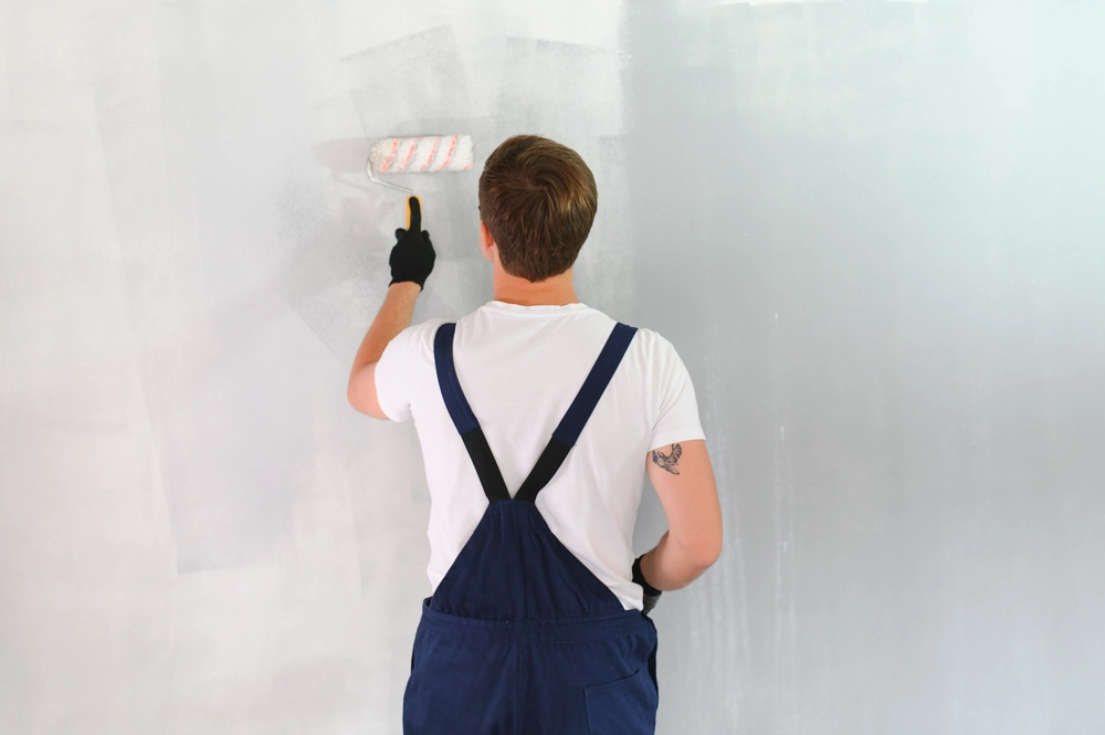 Malowanie ścian: pracownik w ogrodniczkach nanoszący jasną farbę wałkiem na gładką powierzchnię ściany
